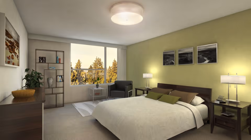 A well-lit bedroom with a large bed featuring white bedding and green and brown pillows. There are two bedside tables with lamps on each side of the bed. Above the bed, three black and white framed pictures are hung on a light green wall. A window shows trees outside, and there is a black armchair next to a small table with a plant. A dresser with a plant and decorative bowl is on the left side of the room, along with a tall shelving unit holding books and decor items.