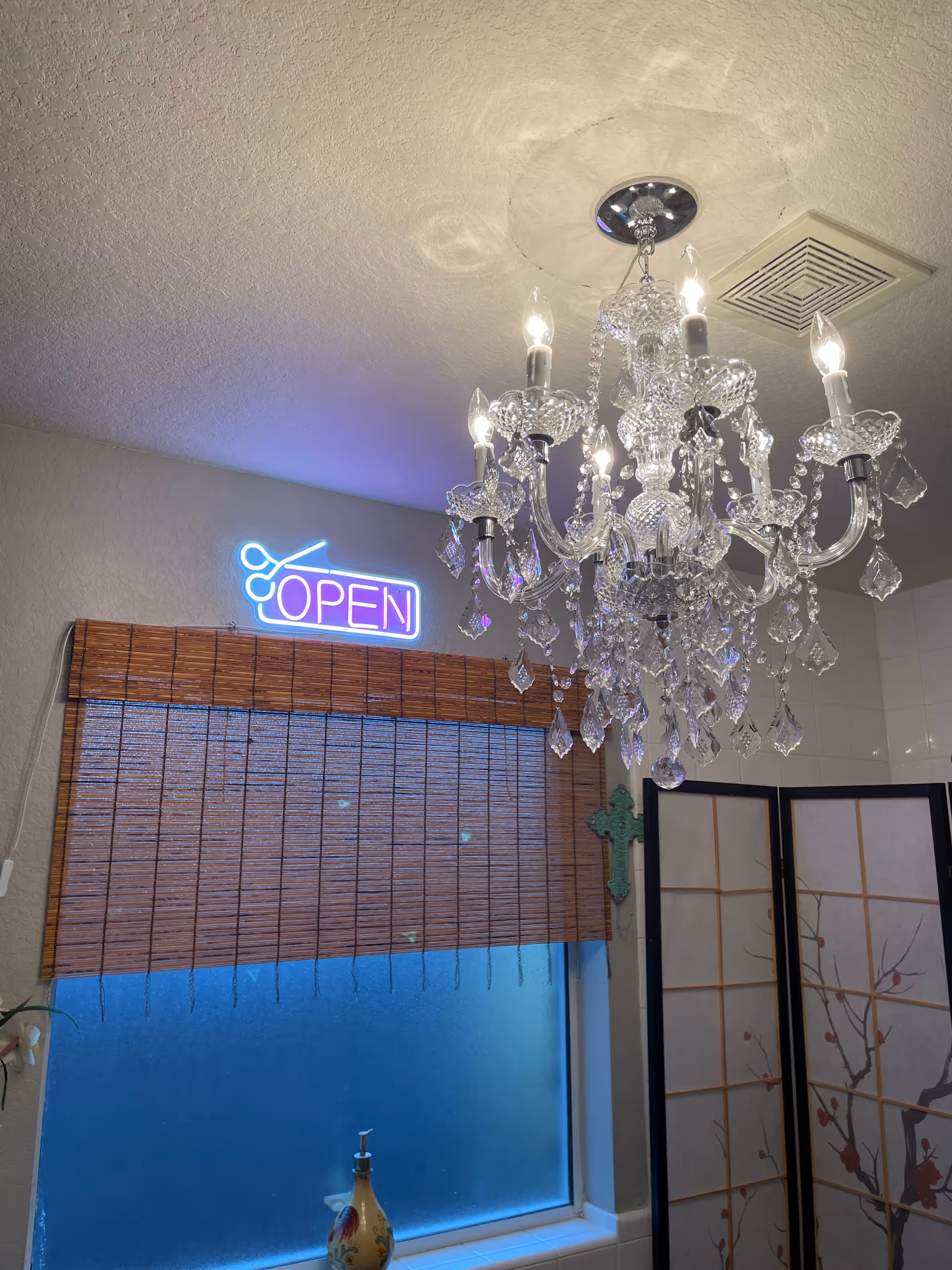 A crystal chandelier hangs over a window with a bamboo blind and a neon "OPEN" sign, next to a decorative folding screen.
