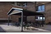 Outdoor patio area with a metal canopy providing shade over several tables and chairs, adjacent to a brick building with multiple windows.