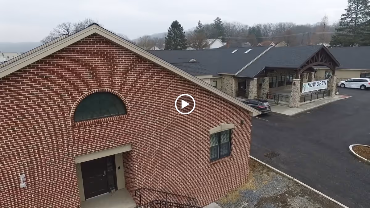 Aerial view of a brick building's front exterior and adjacent entrance canopy with a 'NOW OPEN' banner and parking area.