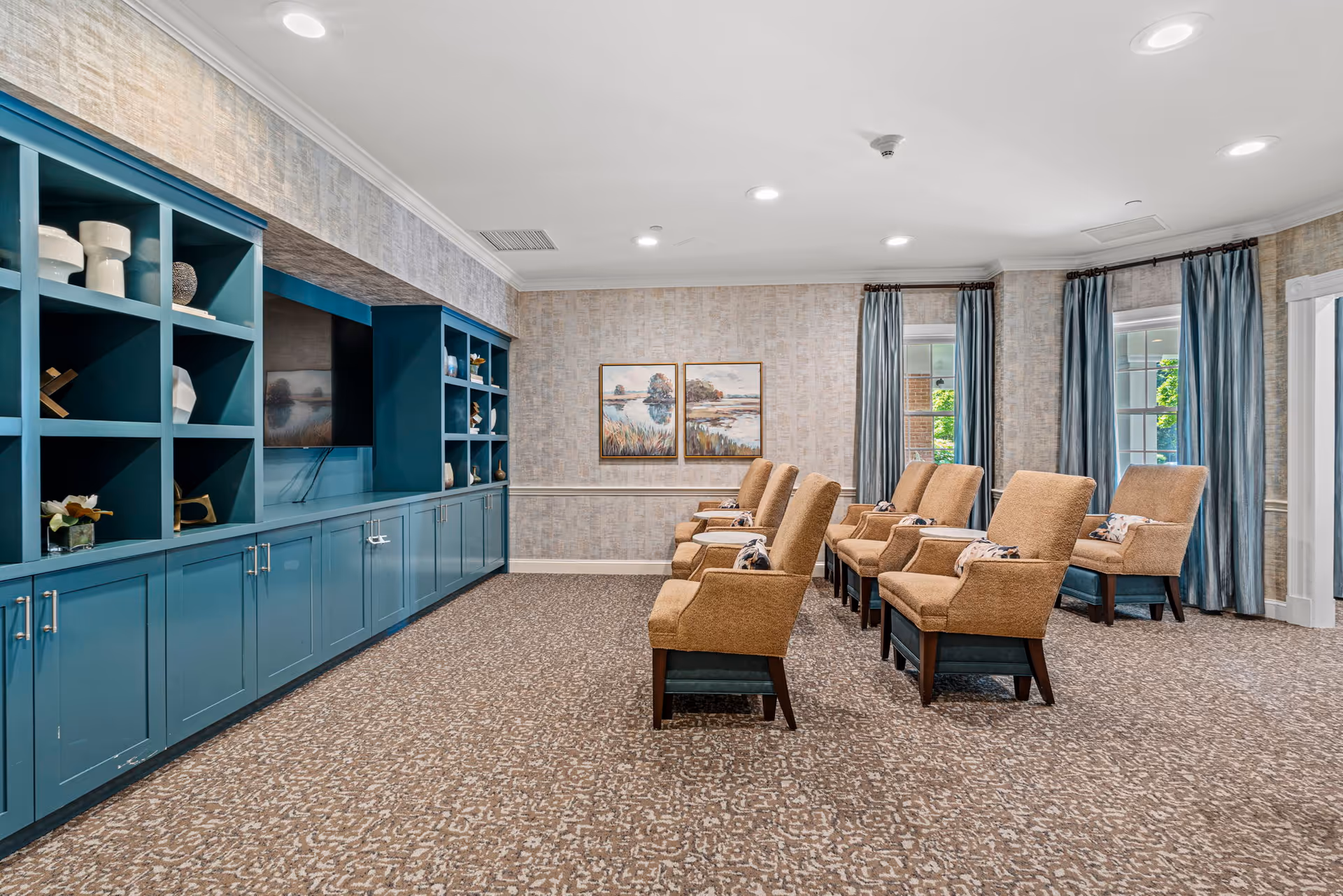 A well-lit room with a row of beige upholstered chairs facing a large built-in blue shelving unit with a mounted flat-screen TV. The room has patterned carpet, light-colored textured wallpaper, two framed landscape paintings on the wall, and windows with blue curtains letting in natural light.