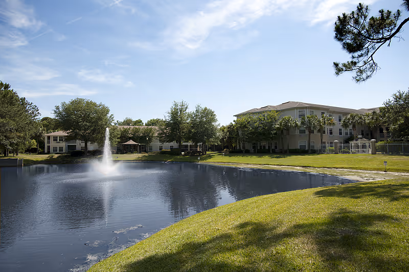 View of a pond with a central fountain in front of multi-story senior living buildings, grassy shore, and trees under a blue sky.