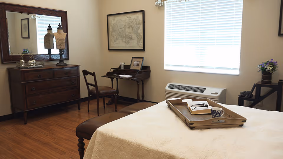 A cozy bedroom with a wooden floor, a bed with a white quilt and a wooden tray holding a book and a cup. There is a wooden dresser with a large mirror above it, a small desk with a chair, a framed map on the wall, and a window with blinds letting in natural light. A small table with a flower arrangement is also visible.