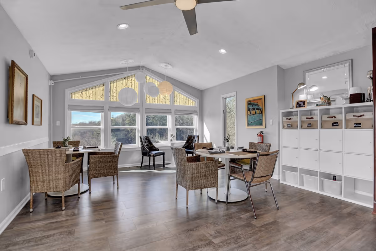 A bright and airy dining area with several tables and chairs arranged for seating. The room features large windows with a scenic outdoor view, hanging paper lanterns from the ceiling, a ceiling fan, and a white storage unit with labeled bins. The walls are decorated with framed artwork and a fire extinguisher is mounted near the door.
