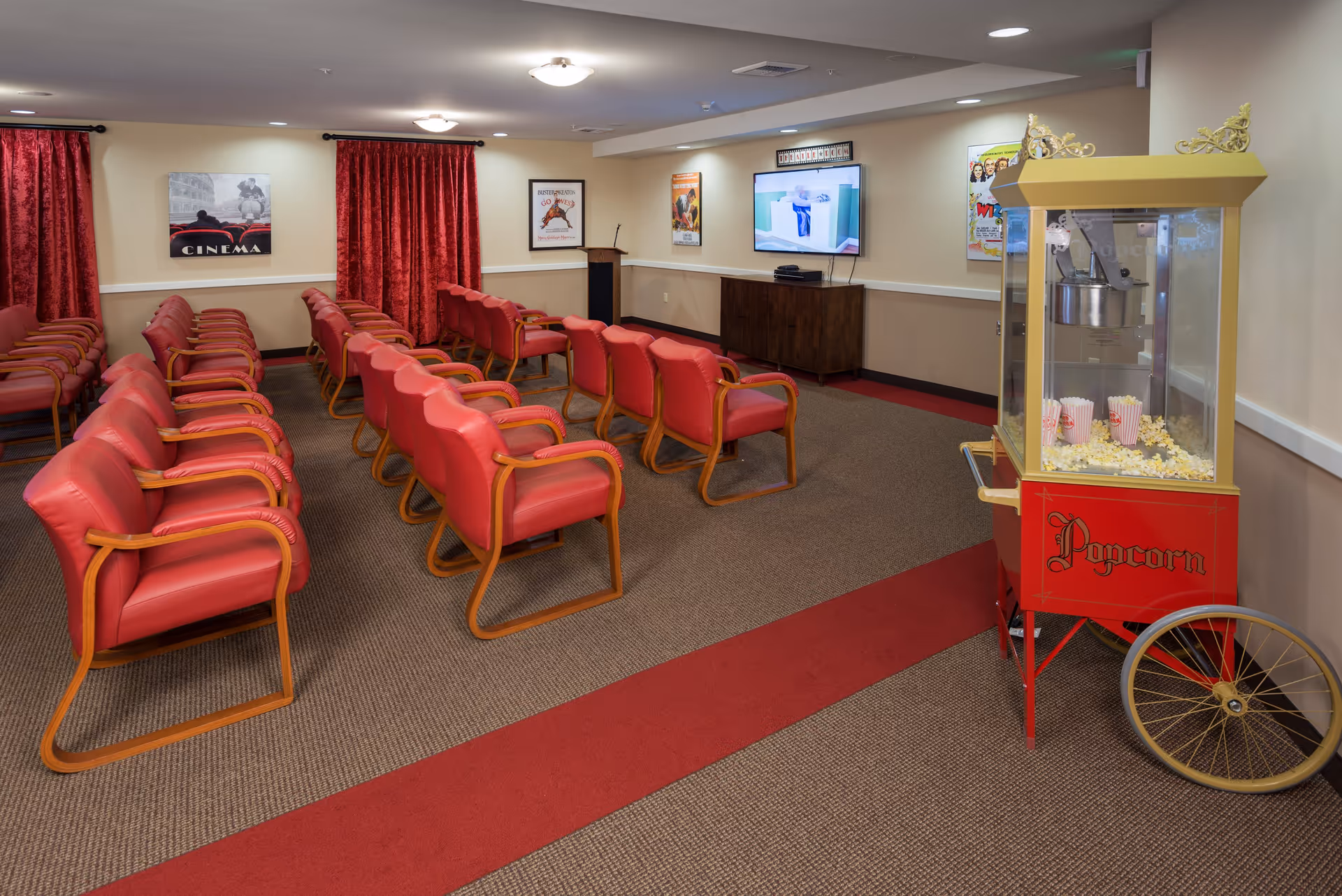 A small movie theater room with rows of red cushioned chairs with wooden armrests facing a wall-mounted TV screen. Red curtains cover the windows, and movie posters decorate the walls. A vintage-style popcorn machine filled with popcorn is positioned on the right side of the room.