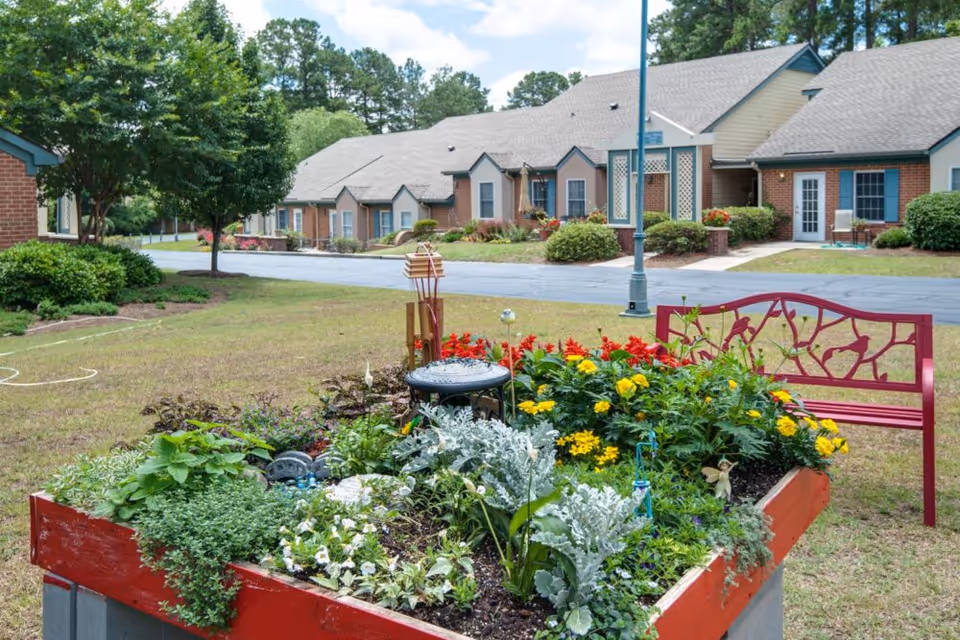 Raised planter filled with colorful flowers and greenery, a red decorative bench, and single-story brick residential buildings and lawn in the background.