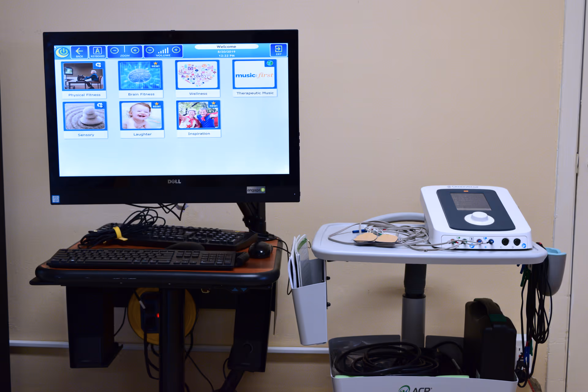 A computer monitor on a desk displaying a wellness program interface with categories like Physical Fitness, Brain Fitness, Wellness, Therapeutic Music, Sensory, Laughter, and Inspiration. Next to the desk is a medical device with wires and electrodes on a small table against a beige wall.