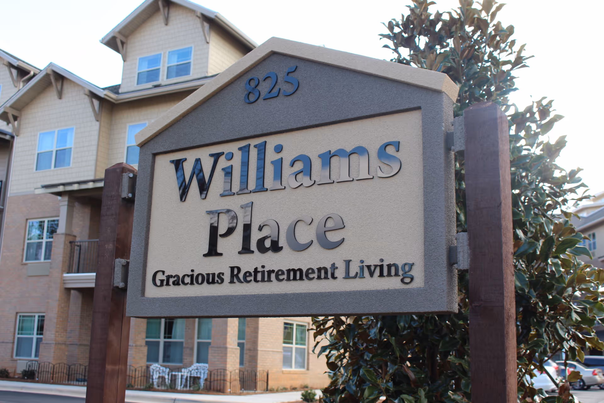 A close-up view of a sign for Williams Place Gracious Retirement Living with the number 825 above it. In the background, there is a multi-story residential building with several windows and balconies, along with some greenery and parked cars.