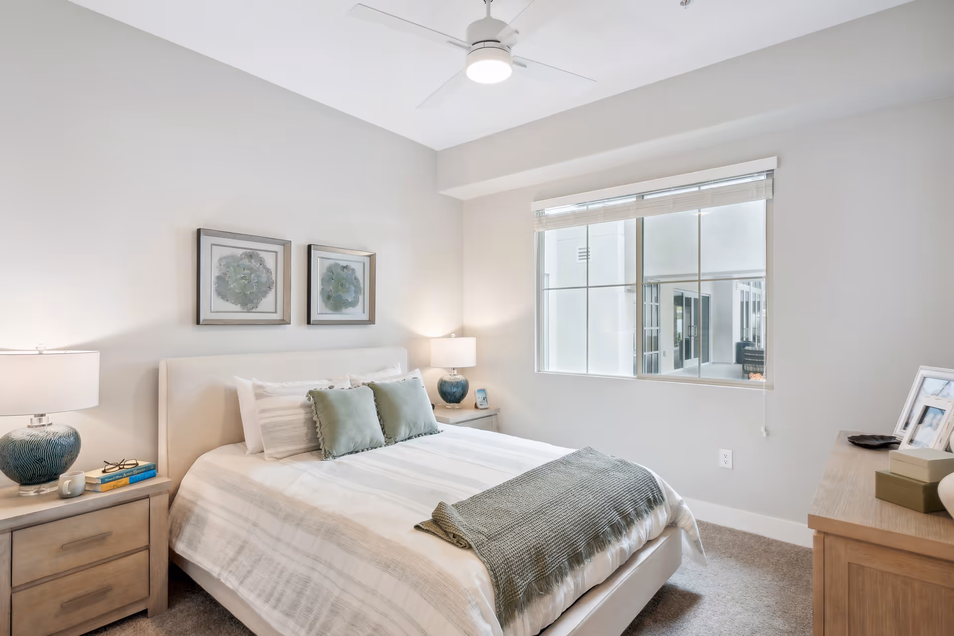 A bright and clean bedroom with a neatly made bed featuring white and light gray striped bedding, two green pillows, and a green throw blanket at the foot. There are two nightstands on either side of the bed, each with a lamp and decorative items. Above the bed are two framed botanical prints. A large window with blinds lets in natural light, and a ceiling fan with a light fixture is mounted on the ceiling.