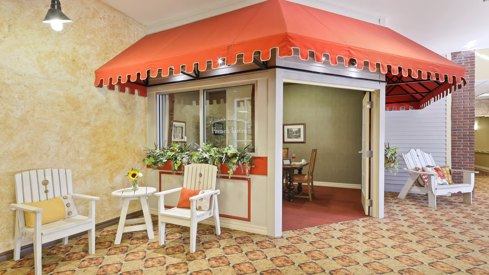 Indoor seating area with two white wooden chairs with cushions and a small round table with a vase holding a sunflower. Adjacent to the seating area is a small room with a red awning labeled 'French bistro' with a table and chairs inside. The floor has a patterned tile design and the walls are textured and painted in warm tones.
