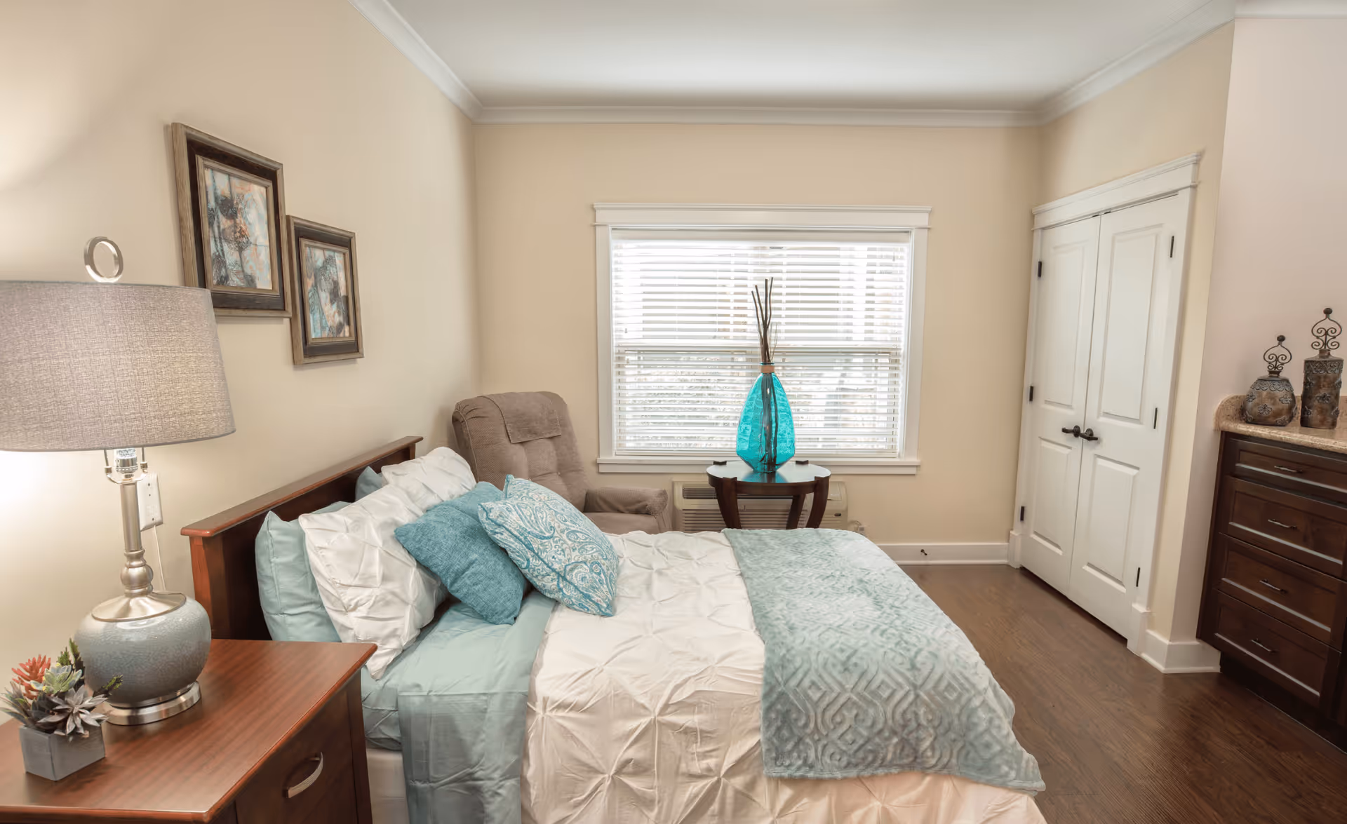 A cozy bedroom with a neatly made bed featuring white and teal bedding and multiple pillows. To the left of the bed is a wooden nightstand with a lamp and a small plant. On the wall above the bed are two framed abstract artworks. In front of the bed is a window with white blinds, and beneath the window is a small wooden table with a large blue decorative vase. To the right is a double-door closet and a wooden dresser with decorative items on top. The room has beige walls and wooden flooring.