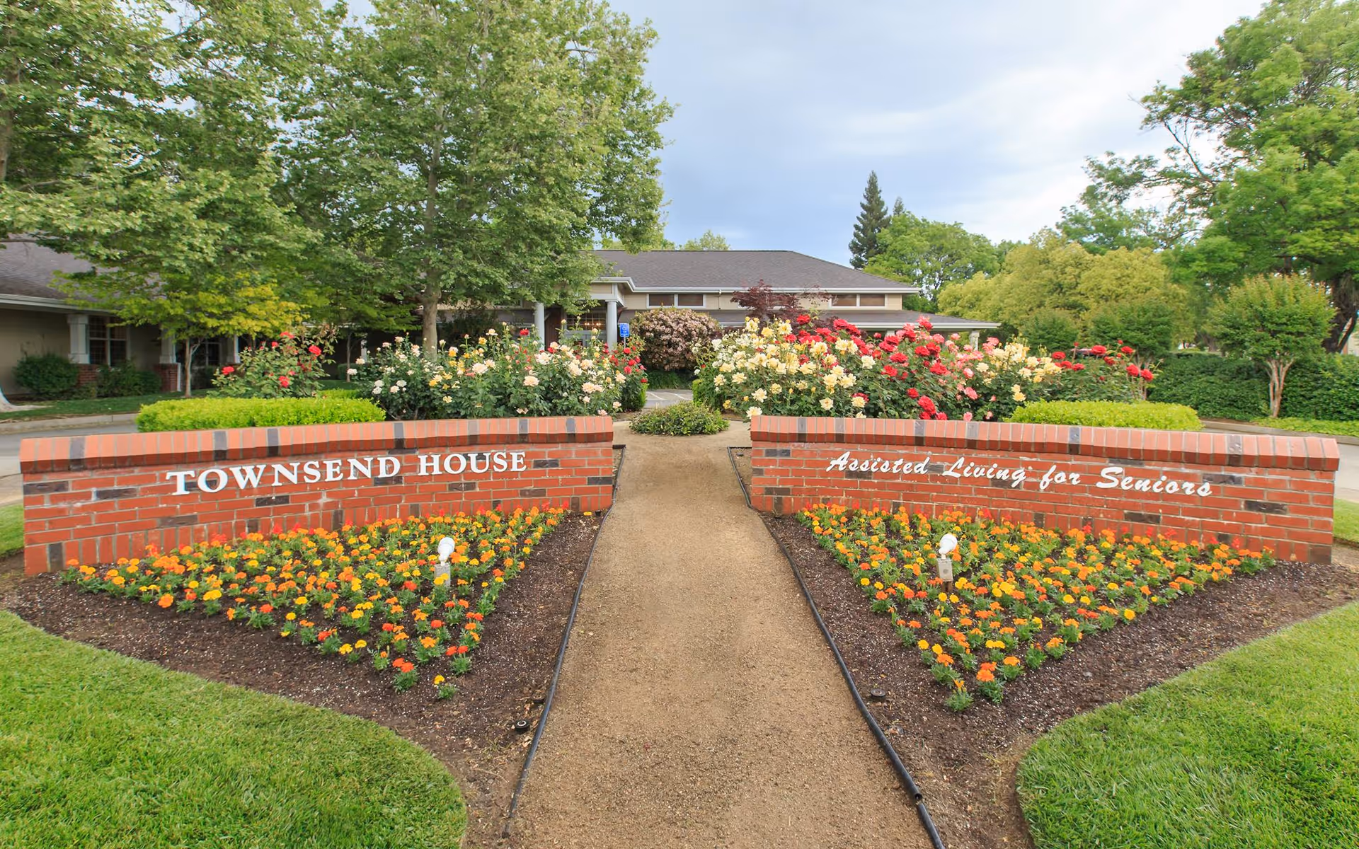 Entrance pathway leading to Townsend House, an assisted living facility for seniors, with neatly maintained flower beds and bushes on either side and a brick sign displaying the facility's name and purpose.