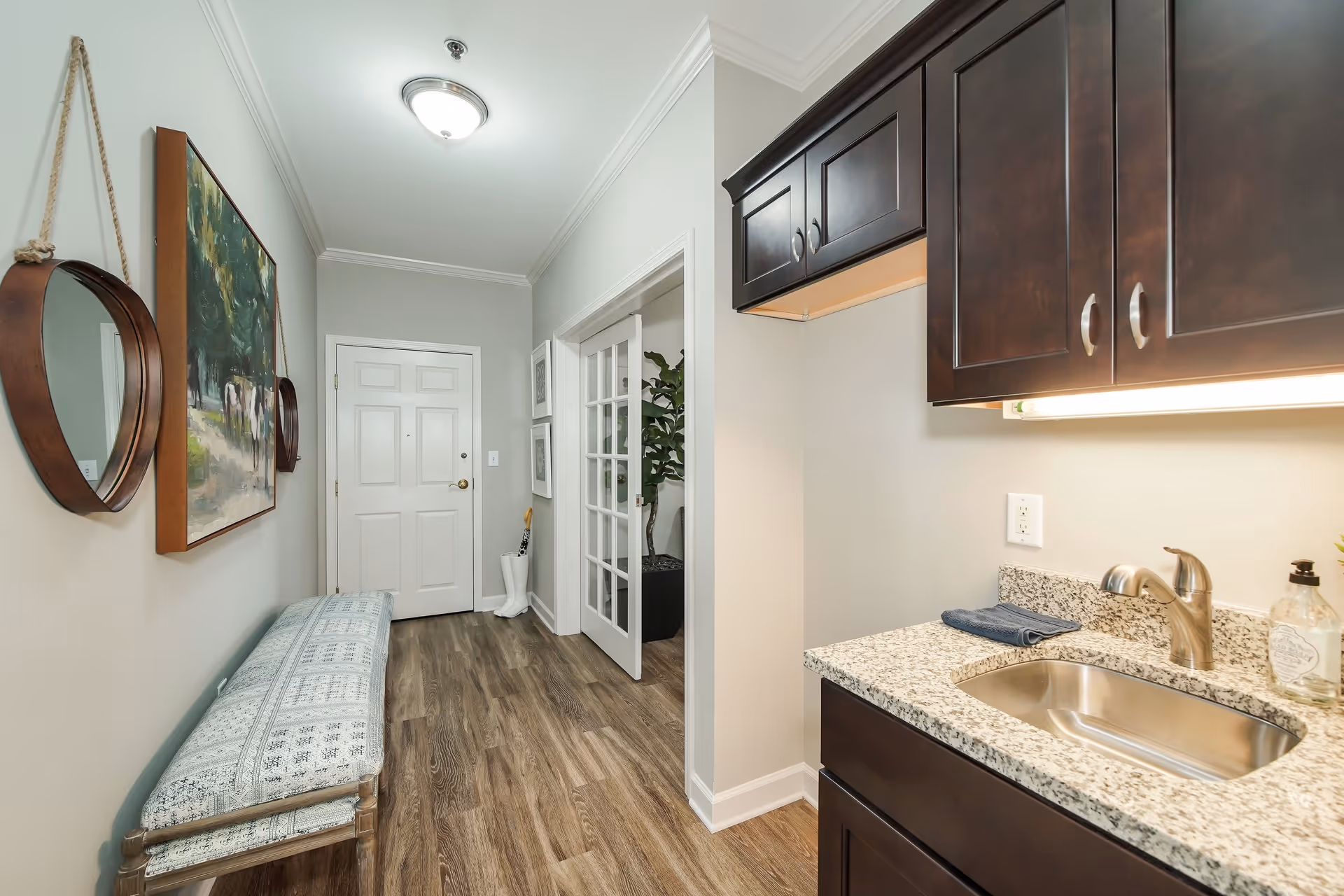 Interior view of a hallway with a white front door at the end. On the left wall, there is a round hanging mirror and a framed painting above a cushioned bench. On the right side, there is a small kitchenette area with dark wood cabinets, a granite countertop, a stainless steel sink, and a faucet. The floor is wood, and there is a ceiling light fixture.