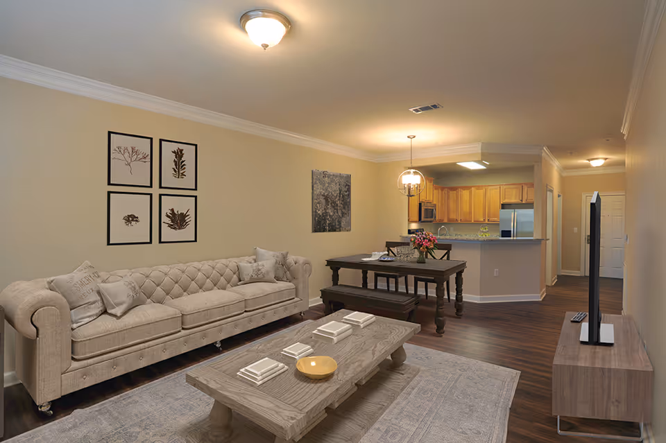 A cozy living room and dining area in an apartment with a beige tufted sofa, a wooden coffee table with books and a bowl, a dining table with chairs and a bench, and a kitchen with wooden cabinets and stainless steel appliances in the background.