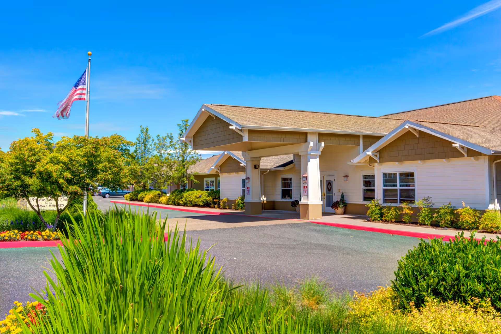 Single-story senior living facility entrance with a covered drop-off, American flag and landscaped grounds under a blue sky.