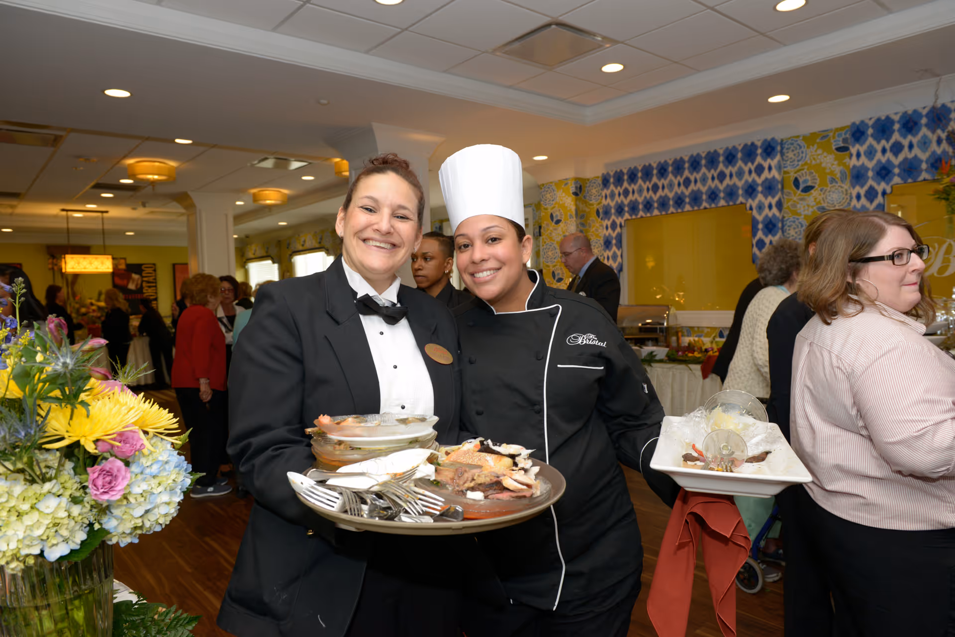 Two smiling staff members at The Bristal Assisted Living at Sayville, one dressed in a black tuxedo with a bow tie holding a tray of dishes and utensils, and the other in a chef's uniform and hat holding a tray with empty dessert glasses. In the background, several people are mingling in a decorated room with floral arrangements and patterned wallpaper.