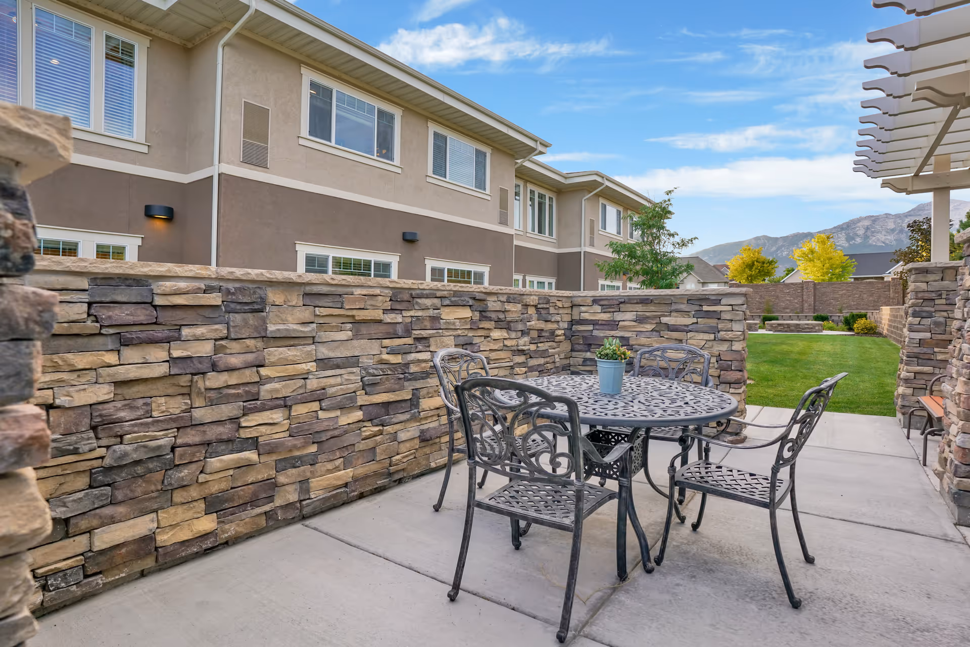 Outdoor patio area with a round metal table and four matching chairs on a concrete floor. The patio is enclosed by a stone wall, with a small potted plant on the table. In the background, there is a grassy area, trees, and a two-story building under a partly cloudy sky.