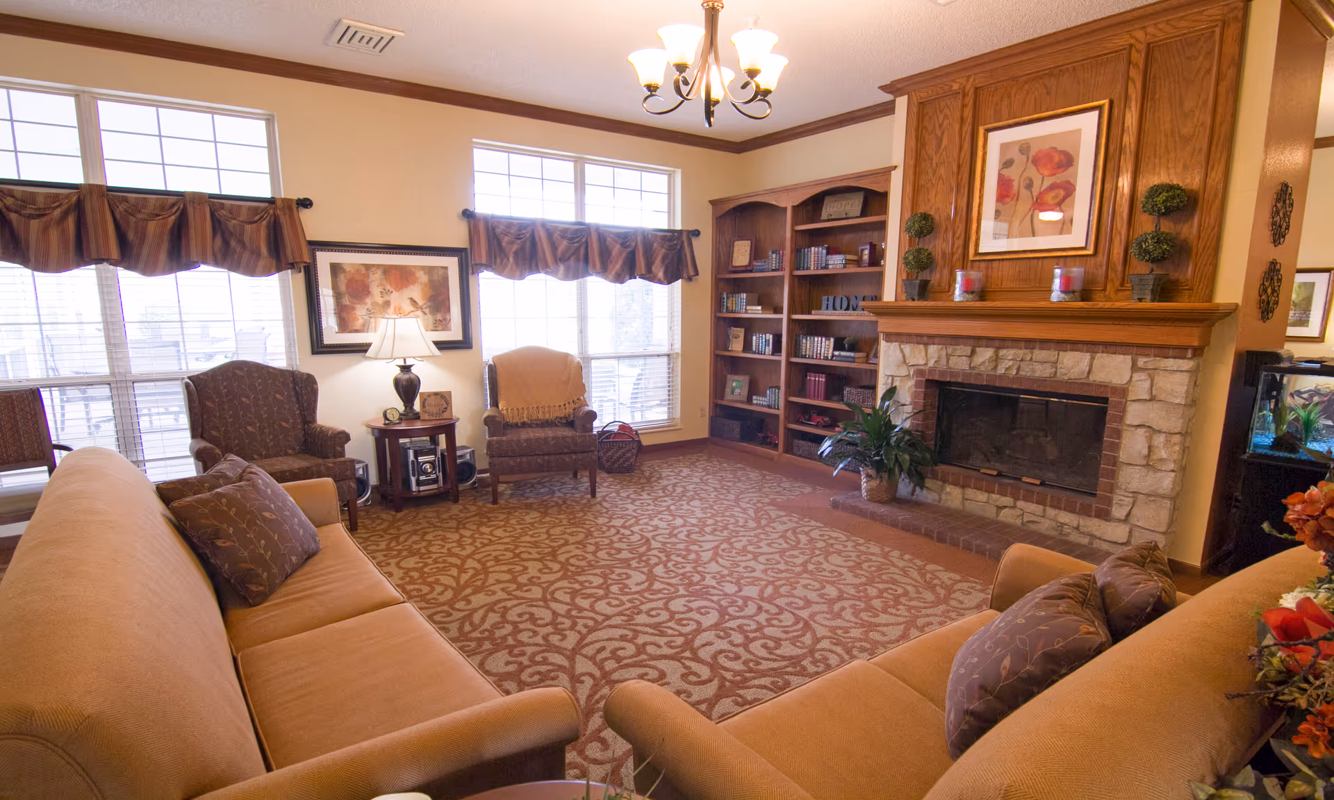 A cozy living room with two beige sofas facing each other, two armchairs, a patterned carpet, a wooden bookshelf filled with books and decorative items, and a stone fireplace with a wooden mantle adorned with plants and candles. Large windows with valances allow natural light to fill the room.