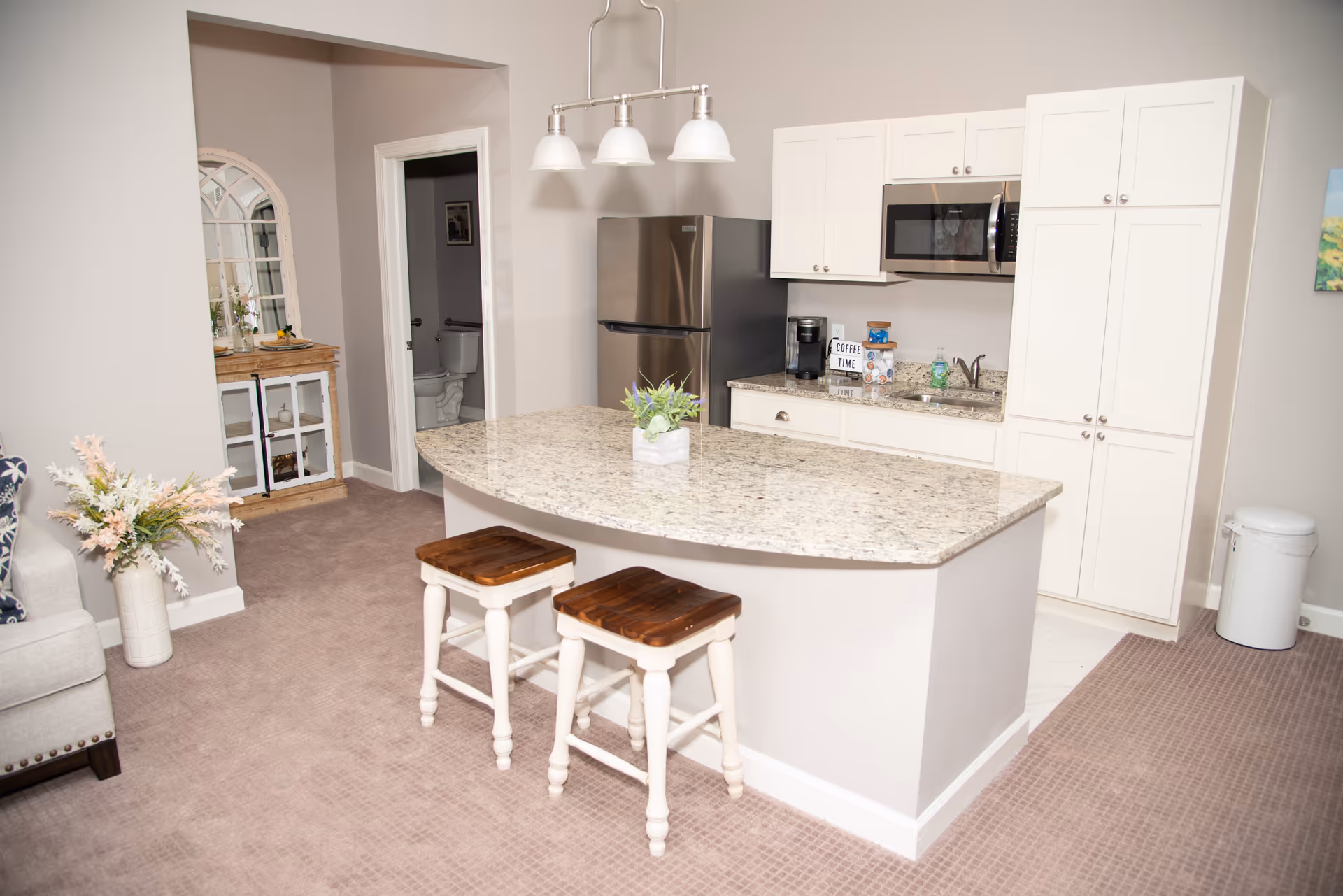 Open kitchen area with a granite island and two wooden stools, stainless steel refrigerator, white cabinets, and pendant lights.