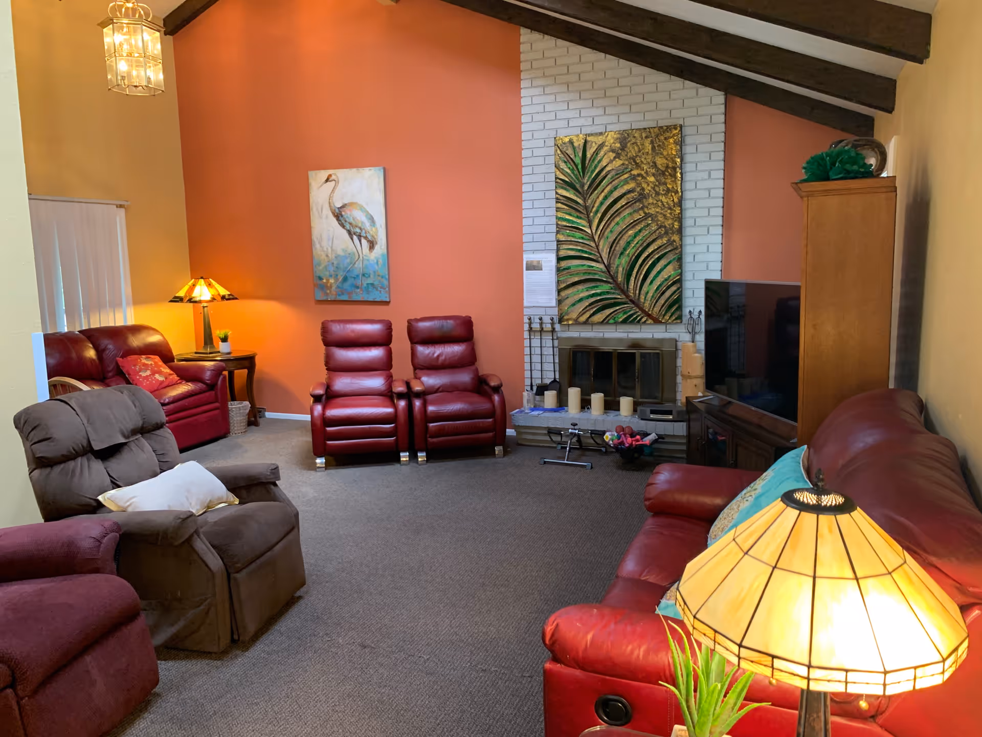 A cozy living room with multiple recliner chairs and sofas in shades of red and brown. The room features a white brick fireplace adorned with candles and a large decorative painting of green leaves. There is a TV on a wooden stand to the right and two paintings on the walls, one of a bird and another with green leaves. The walls are painted in warm tones of orange and yellow, and the ceiling has exposed dark wooden beams. Two table lamps with stained glass shades provide warm lighting.
