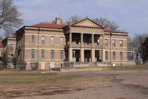 Two-story brick building with columns and a central pedimented entrance, set on a dirt foreground.