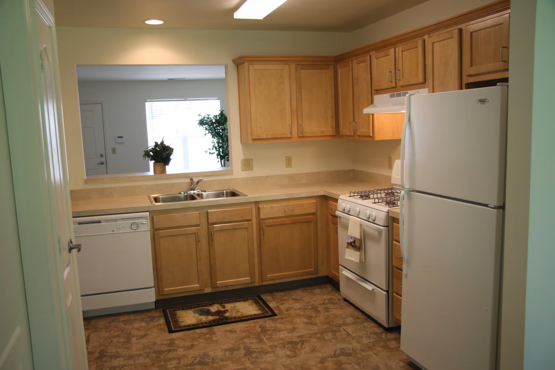 A clean kitchen with light wood cabinets, a white refrigerator, a white gas stove with a towel hanging on the handle, a double sink, and a dishwasher. There is a pass-through window with a view into a living area with plants and a window letting in natural light.