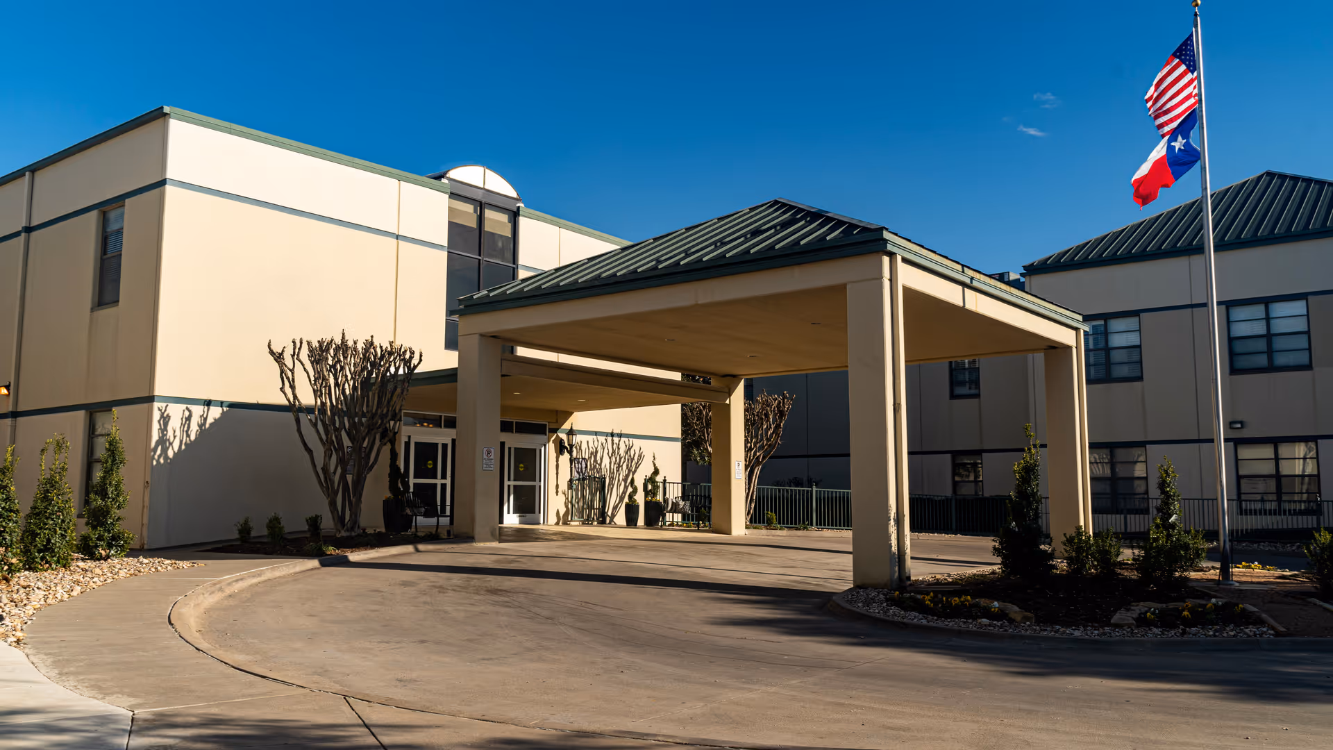 Exterior view of Cityview Nursing and Rehabilitation Center showing the main entrance with a covered drop-off area, beige building walls, trimmed bushes, and flagpoles with American and Texas flags against a clear blue sky.