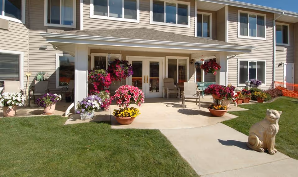 Outdoor patio area of a senior living facility with a concrete walkway, green lawn, multiple flower pots with colorful flowers, hanging flower baskets, patio chairs, and a cat statue on the grass near the walkway. The building exterior is beige with multiple windows and a covered porch.