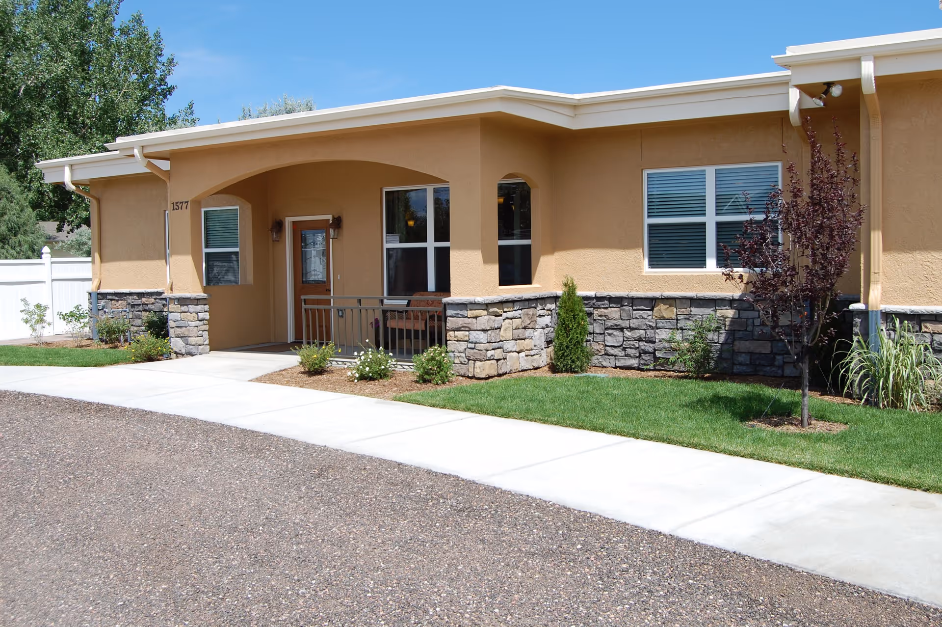 Exterior view of a single-story assisted living facility building with beige stucco walls and stone accents. The entrance features a covered porch with a wooden door and windows, surrounded by small shrubs and a well-maintained lawn. A concrete sidewalk runs in front of the building under a clear blue sky.
