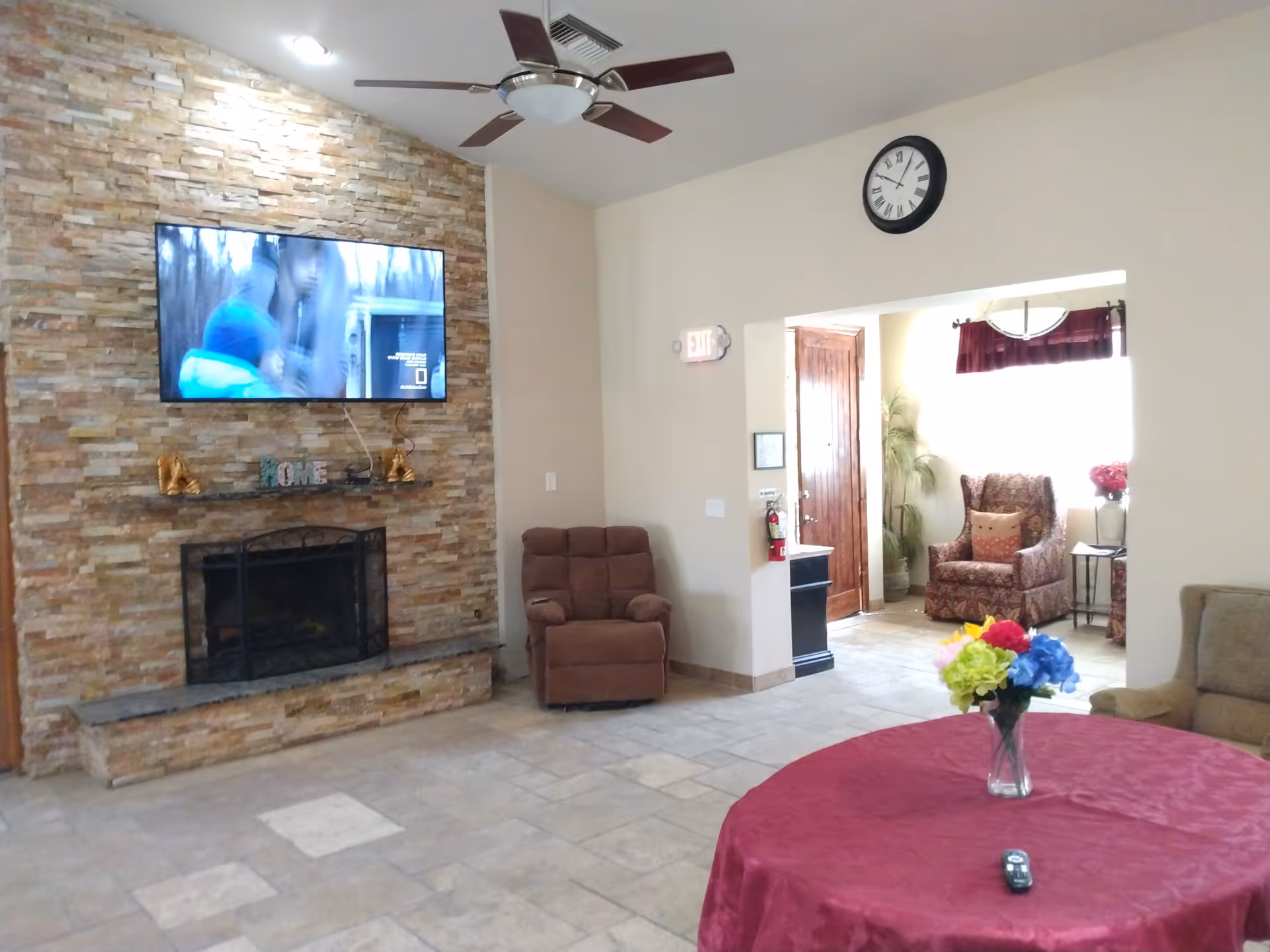A cozy living room area with a stone fireplace and a mounted flat-screen TV above it. There is a brown recliner chair near the fireplace, a round table covered with a red tablecloth and a vase of colorful flowers, and a beige sofa partially visible. A ceiling fan with wooden blades is mounted on the ceiling. In the background, there is an open doorway leading to another room with an armchair, a side table, and a window with curtains. A clock is mounted on the wall above the doorway.