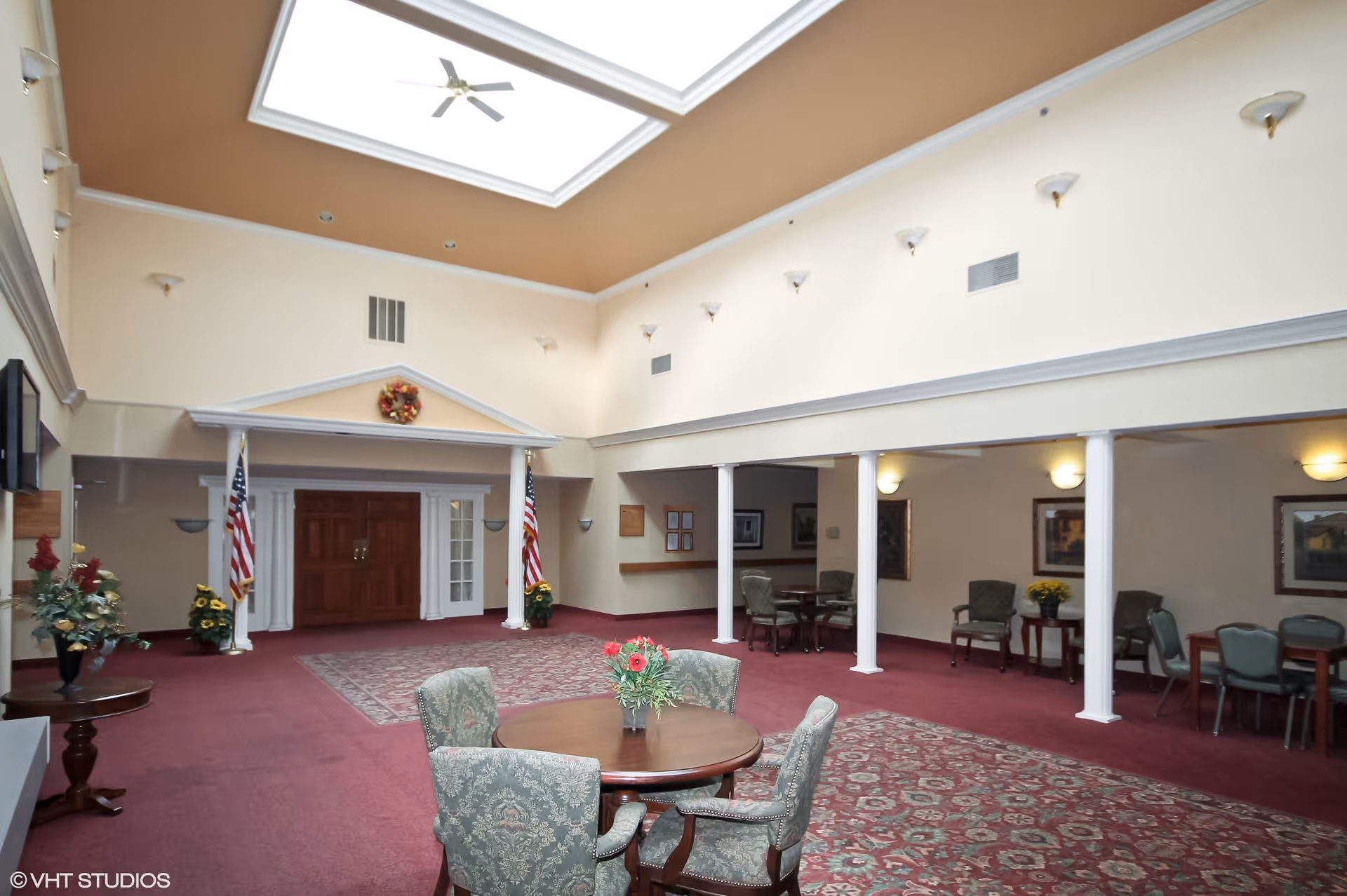 Spacious interior common area with high ceilings and a large skylight. The room features a maroon carpet with patterned rugs, several round tables with upholstered chairs, decorative plants, and American flags flanking a set of double wooden doors. The walls are light-colored with sconces and framed artwork, and white columns separate seating areas.