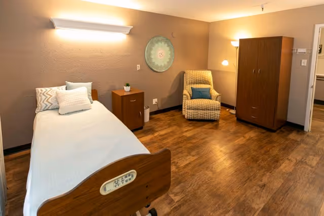 Small furnished senior-care bedroom with a single hospital-style bed, bedside table, upholstered armchair, wardrobe, and warm lighting.