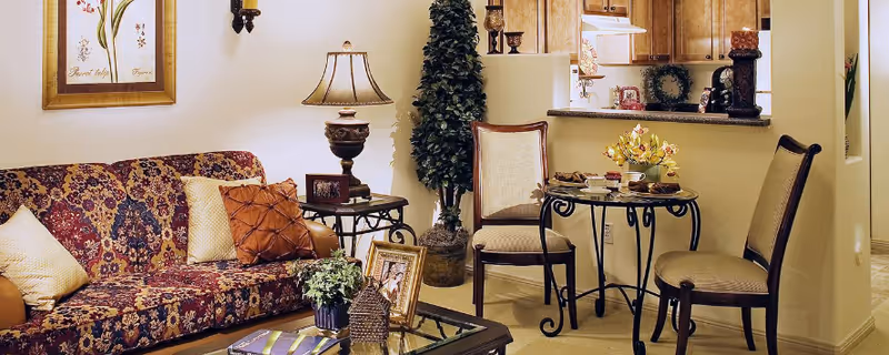 A cozy living room area with a patterned sofa adorned with multiple pillows, a glass coffee table with decorative items, a side table with a lamp, and a small dining area with a round table and two chairs. The background shows a kitchen with wooden cabinets and a countertop.