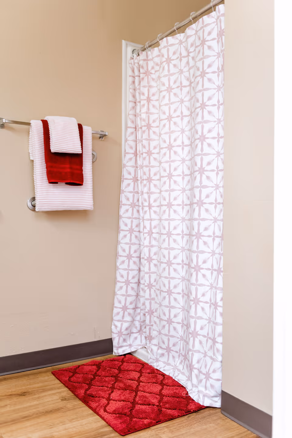 A bathroom corner featuring a shower with a white and light pink patterned shower curtain. Next to the shower, there is a towel rack holding two white towels and one red towel. A red bath mat is placed on the wooden floor in front of the shower.