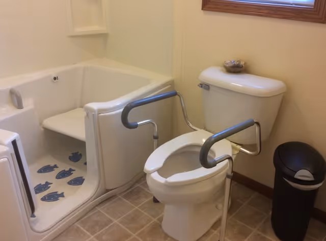 A bathroom with a walk-in bathtub featuring a built-in seat and fish decals on the floor, a toilet equipped with safety grab bars on both sides, a small bowl on top of the toilet tank, and a black trash can next to the toilet. There is a window with a wooden frame above the toilet.