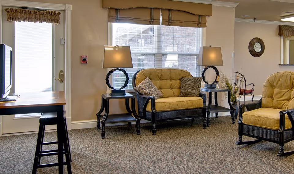 A cozy living room area with a cushioned loveseat and armchair in a wicker style, both with mustard yellow cushions. There are two side tables with matching lamps featuring circular bases. A window with blinds and a valance is behind the loveseat. A clock hangs on the wall, and a desk with a stool is positioned near a door with a window.