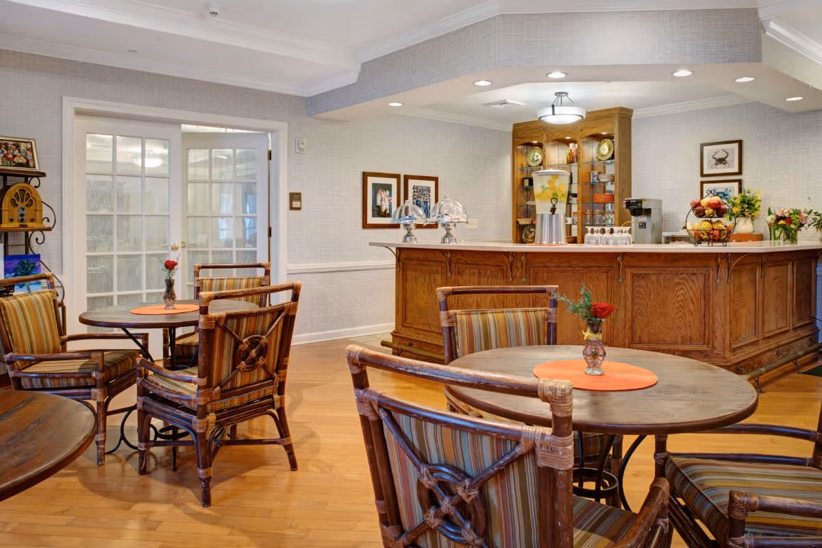 A cozy dining area in a senior living facility with wooden tables and striped cushioned chairs. Each table has a small vase with a single red rose. In the background, there is a wooden counter with a beverage dispenser, coffee machine, and fruit baskets. The walls are light-colored with framed pictures, and there are glass double doors on the left side.