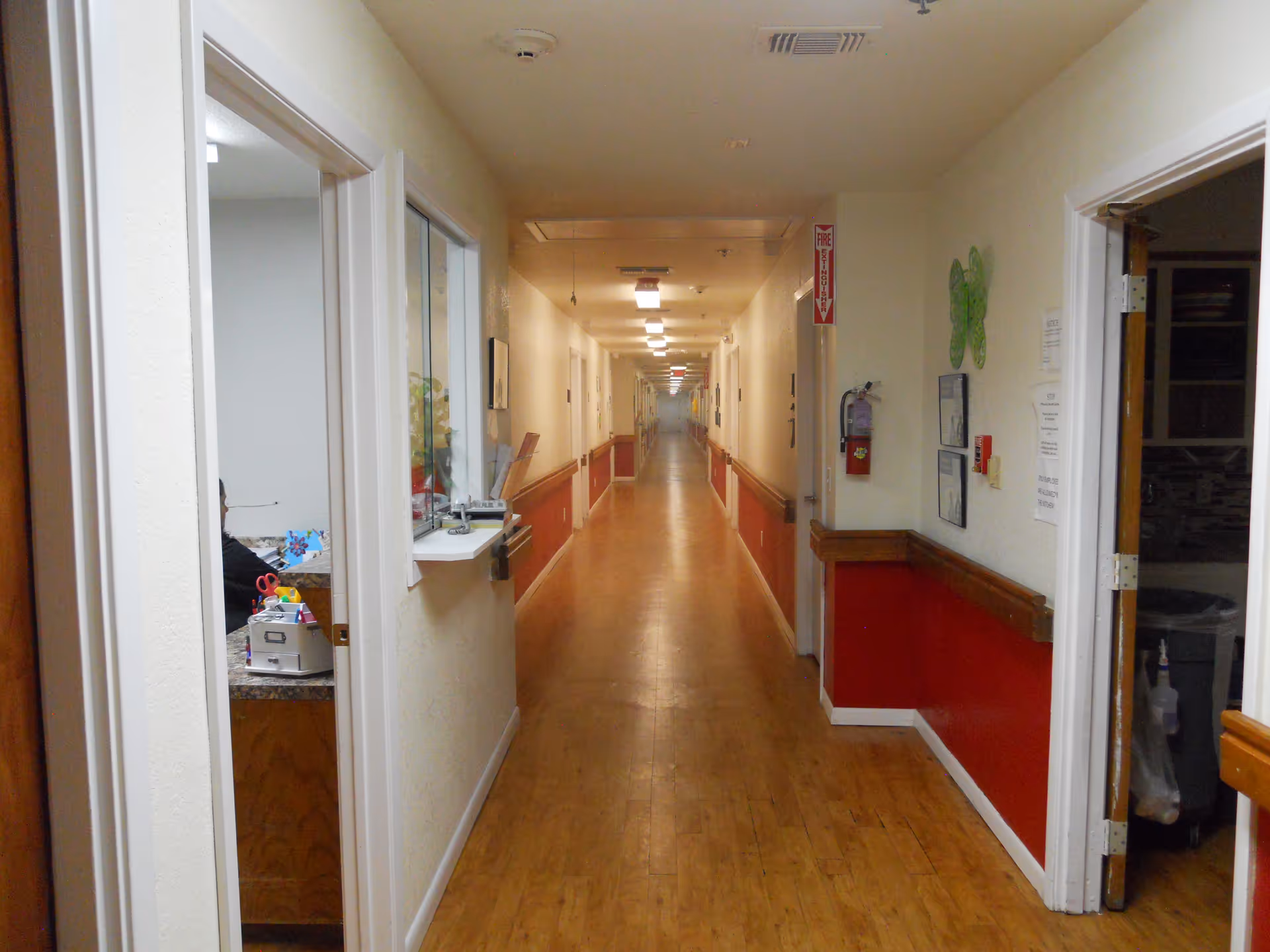 Long indoor hallway with wooden flooring and cream-colored walls with red lower panels. Doors and windows line both sides of the hallway. A fire extinguisher is mounted on the right wall, and fluorescent lights illuminate the corridor ceiling.