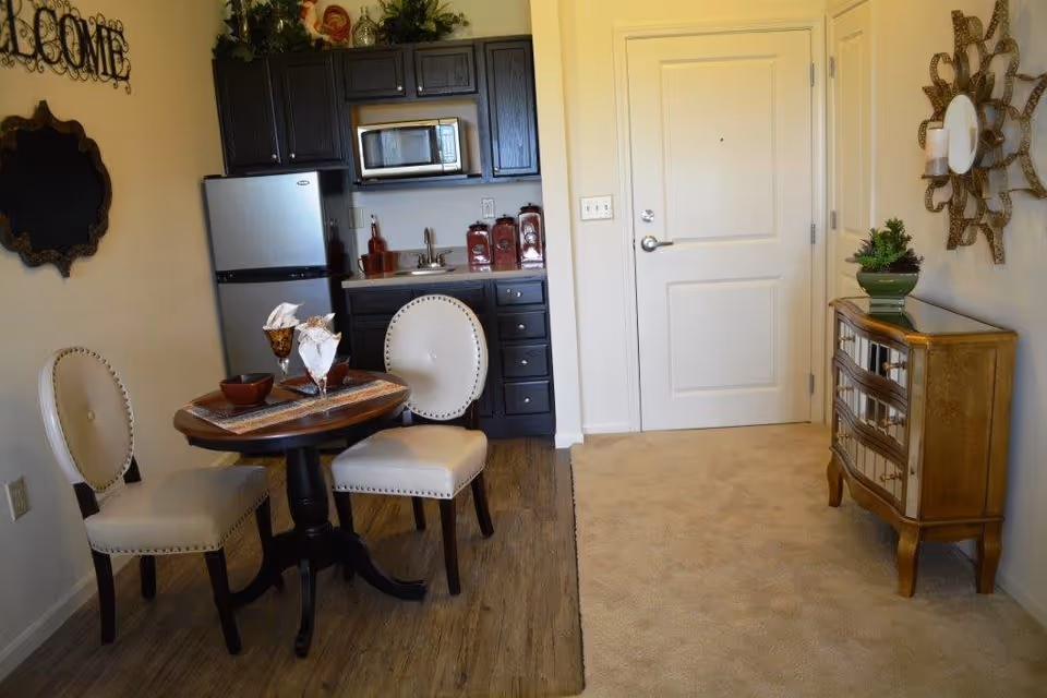 Small dining area with a round wooden table set for two with white upholstered chairs. Behind the table is a kitchenette with dark cabinets, a small refrigerator, microwave, and countertop with a sink. To the right is a white door and a decorative wooden cabinet with a plant on top. The floor transitions from wood to carpet near the door. Wall decorations include a 'WELCOME' sign and a decorative mirror.