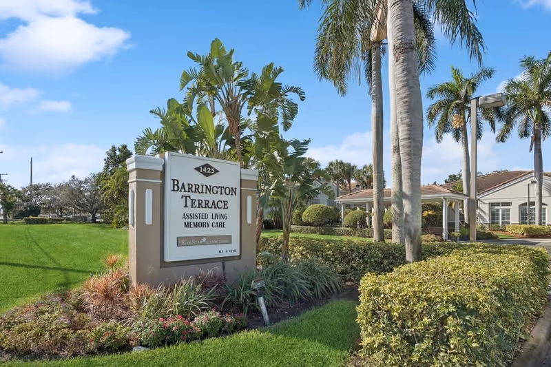 Outdoor view of Barrington Terrace assisted living and memory care facility sign surrounded by green grass, plants, and palm trees under a blue sky with some clouds.