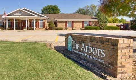 Exterior view of The Arbors at Glendale Gardens senior living facility showing a single-story brick building with a covered entrance, an American flag, and a brick sign with the facility's name in the foreground.