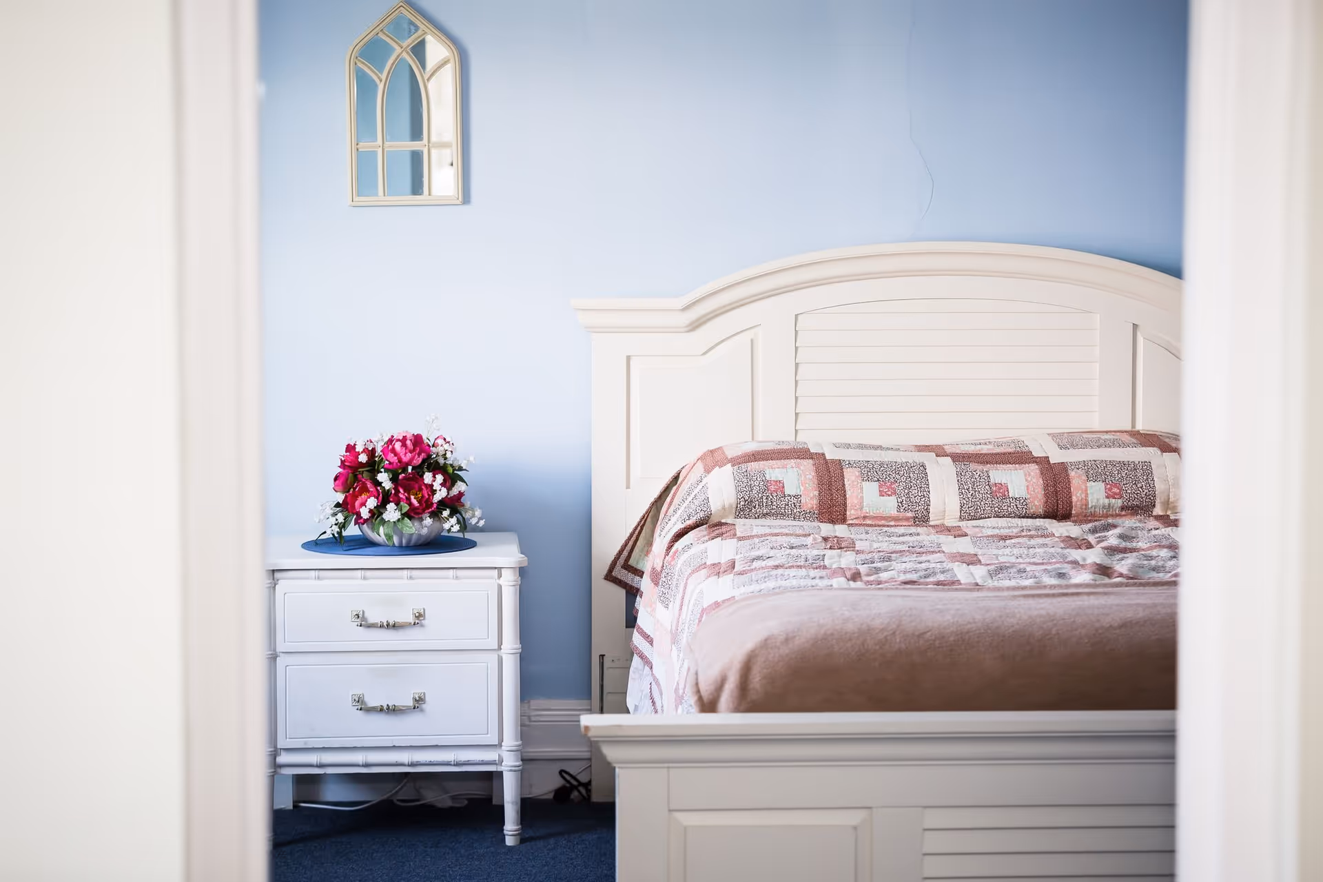 A cozy bedroom with a white wooden bed frame and a patchwork quilt in shades of pink and brown. Next to the bed is a white nightstand with two drawers, topped with a vase of vibrant pink and white flowers. The wall behind the bed is painted light blue and features a decorative arched mirror.