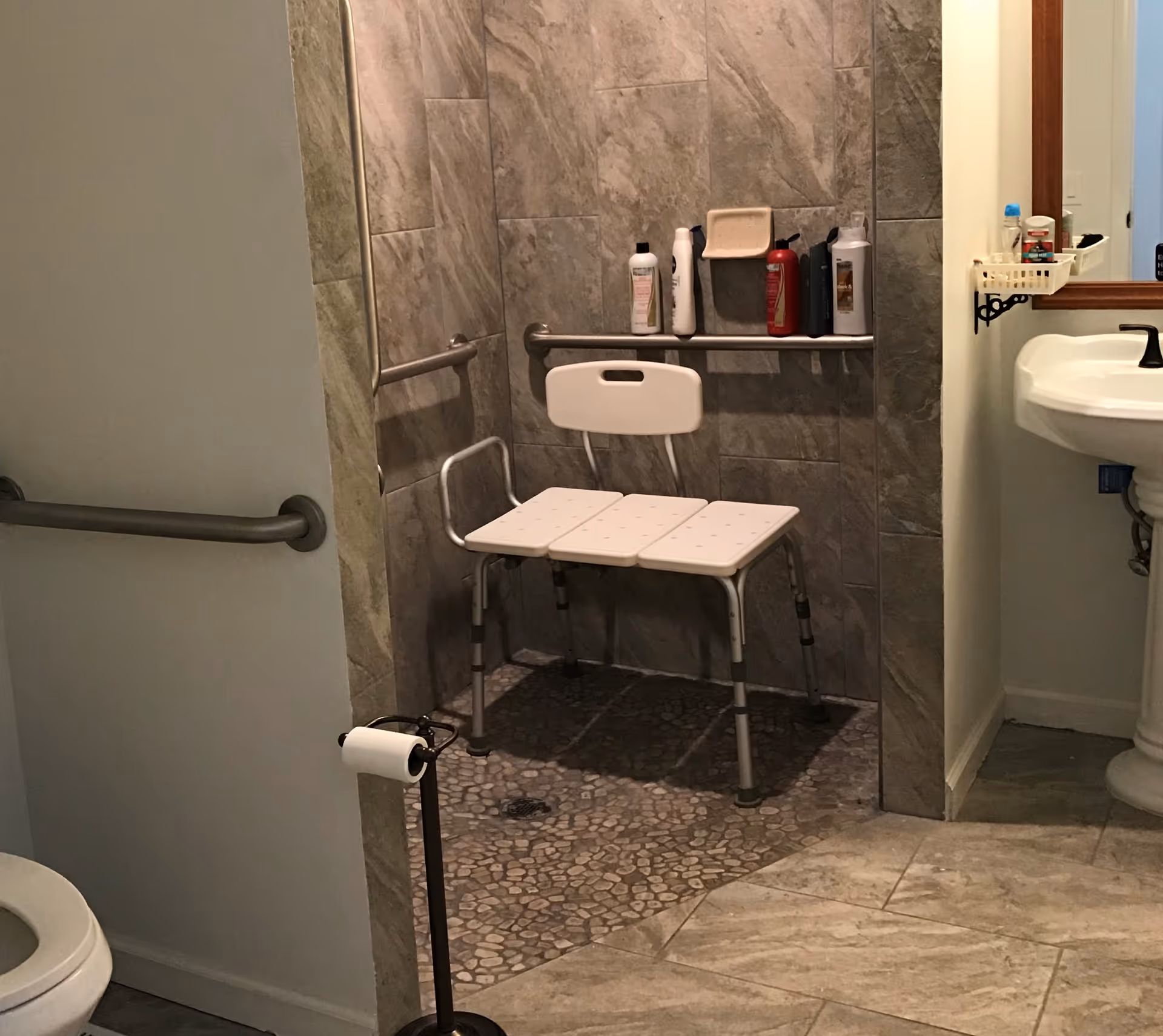 Bathroom with a walk-in shower featuring a white shower chair, grab bars, and various bottles on a built-in shelf. There is a pedestal sink with a mirror and a small basket holding toiletries, a toilet partially visible, and a freestanding toilet paper holder.