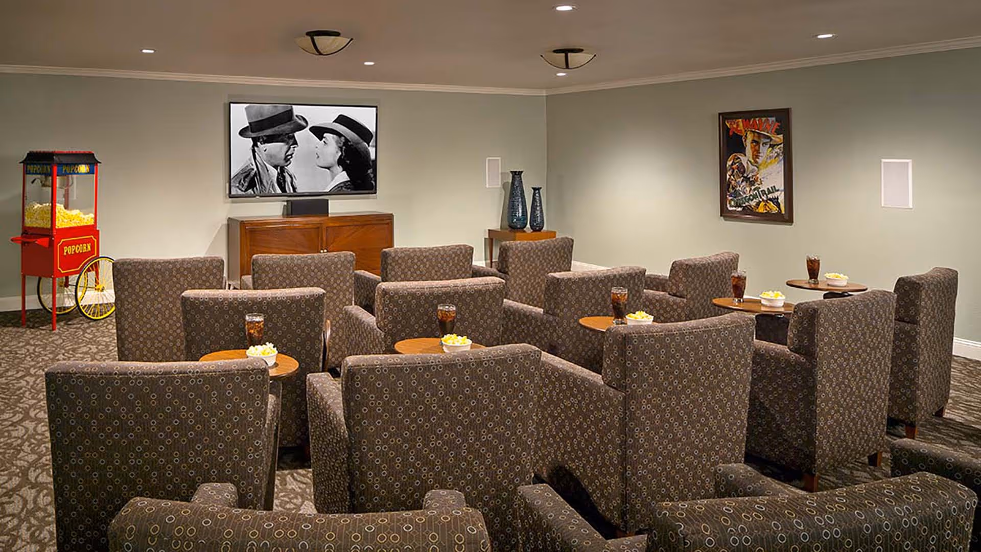 A small movie theater room with multiple patterned armchairs arranged in rows facing a wall-mounted TV screen showing a black and white movie. Small round tables between chairs hold popcorn bowls and glasses of soda. A red popcorn machine is visible in the corner, and framed movie posters hang on the walls.