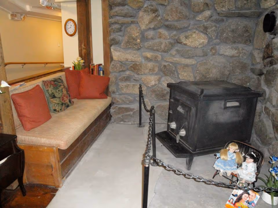 A cozy interior corner with a stone wall and a black wood stove enclosed by a chain barrier. To the left, there is a wooden bench with beige cushions and several red and floral pillows. A small black side table is partially visible. Two dolls are seated on a small chair near the stove, and a clock is mounted on the wall in the background.