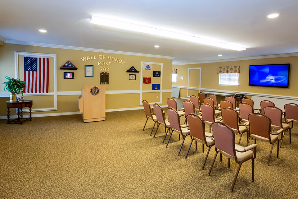 A meeting or presentation room with rows of chairs facing a podium and a wall labeled 'Wall of Honor Post 208' decorated with flags, plaques, and framed items. There is an American flag displayed on the left wall and a flat-screen TV mounted on the right wall. The room has beige walls, carpeted floor, and ceiling lights.