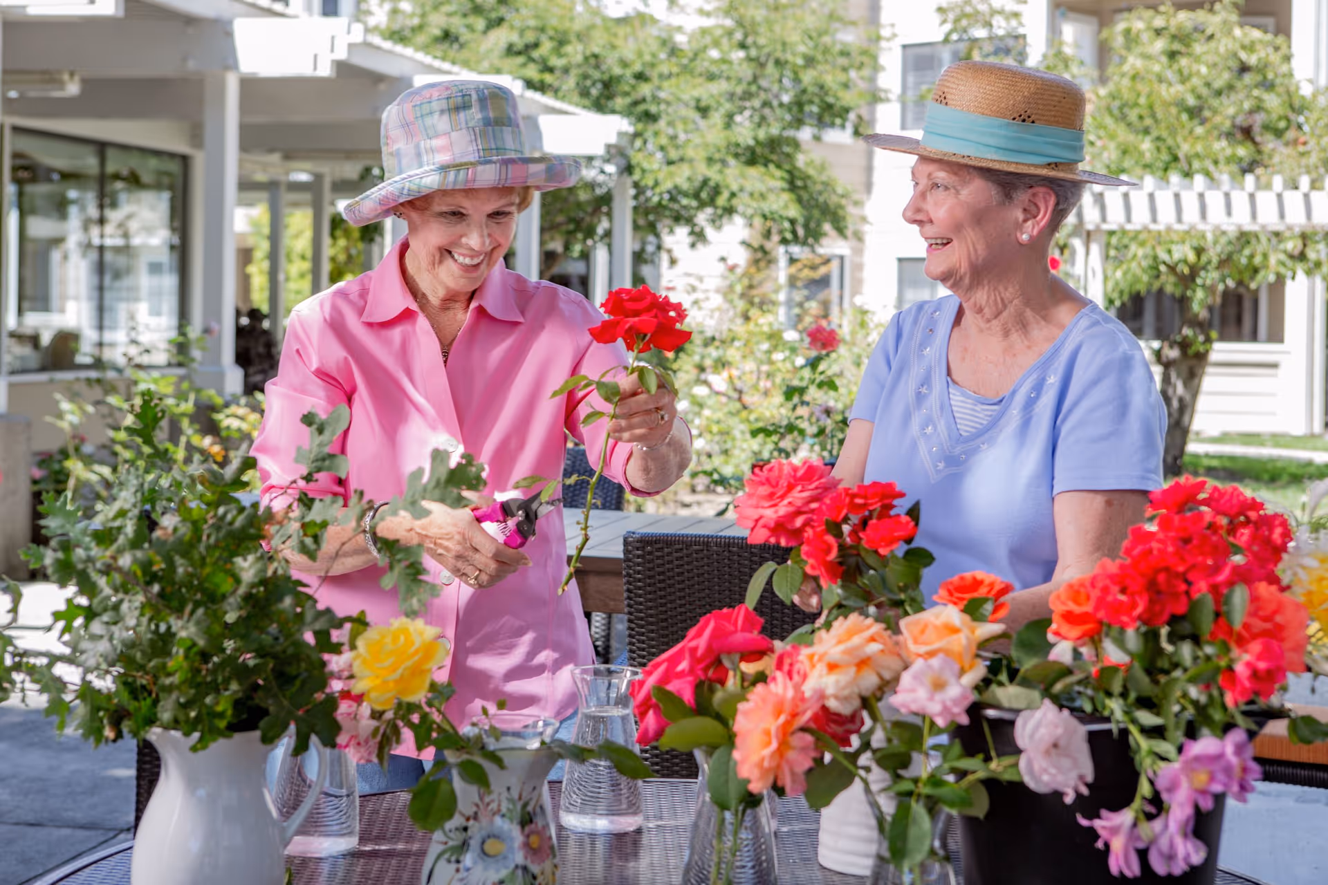 Two elderly women wearing hats are arranging colorful flowers on a table outdoors in a garden area of a senior living facility. One woman in a pink shirt is holding a red rose and pruning shears, while the other woman in a light blue shirt smiles at her. Various vases filled with vibrant flowers are on the table, and greenery and buildings are visible in the background.