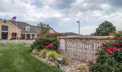 Exterior view of Waldron Place senior living with entrance, sign, lawn, and flowering bushes.