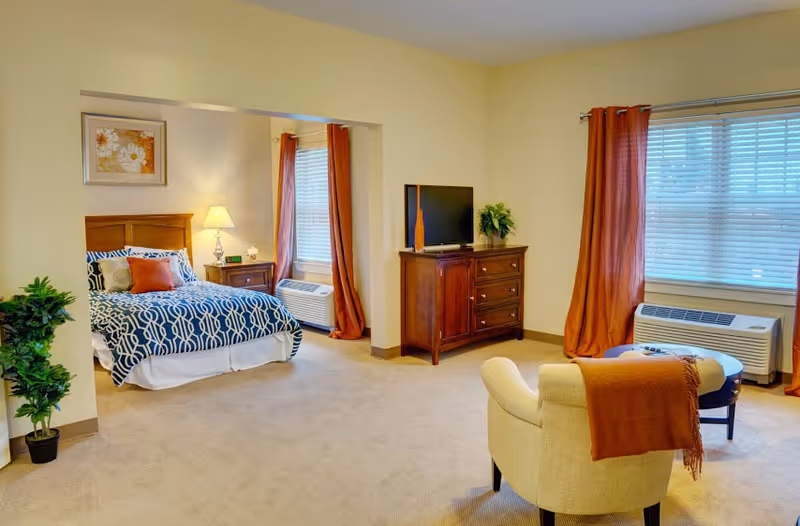 Spacious studio apartment interior with a bed in an alcove, dresser with TV, armchair, and windows with orange curtains.