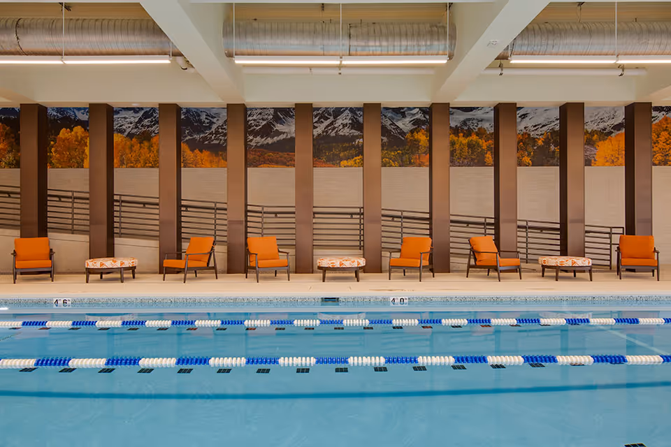Indoor swimming pool with lane dividers and a row of orange lounge chairs in front of a scenic mural.